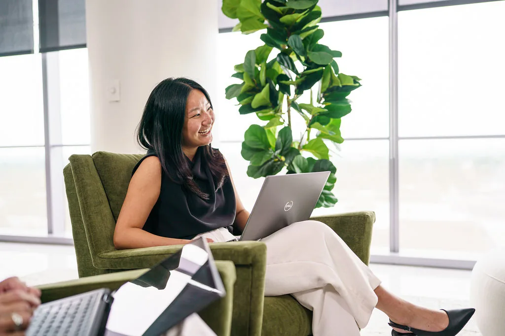 NISA team member smiling while working on a laptop in a green armchair in the sunlit office lounge